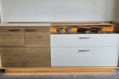 Sideboard in Eiche massiv, satin gebeizt, Absetzung in Lack weiß. Inklusive LED-Beleuchtung am Sockel und im Glasteil. Maß: B 196 x H 83 x T 45 cm. Ohne Deko.